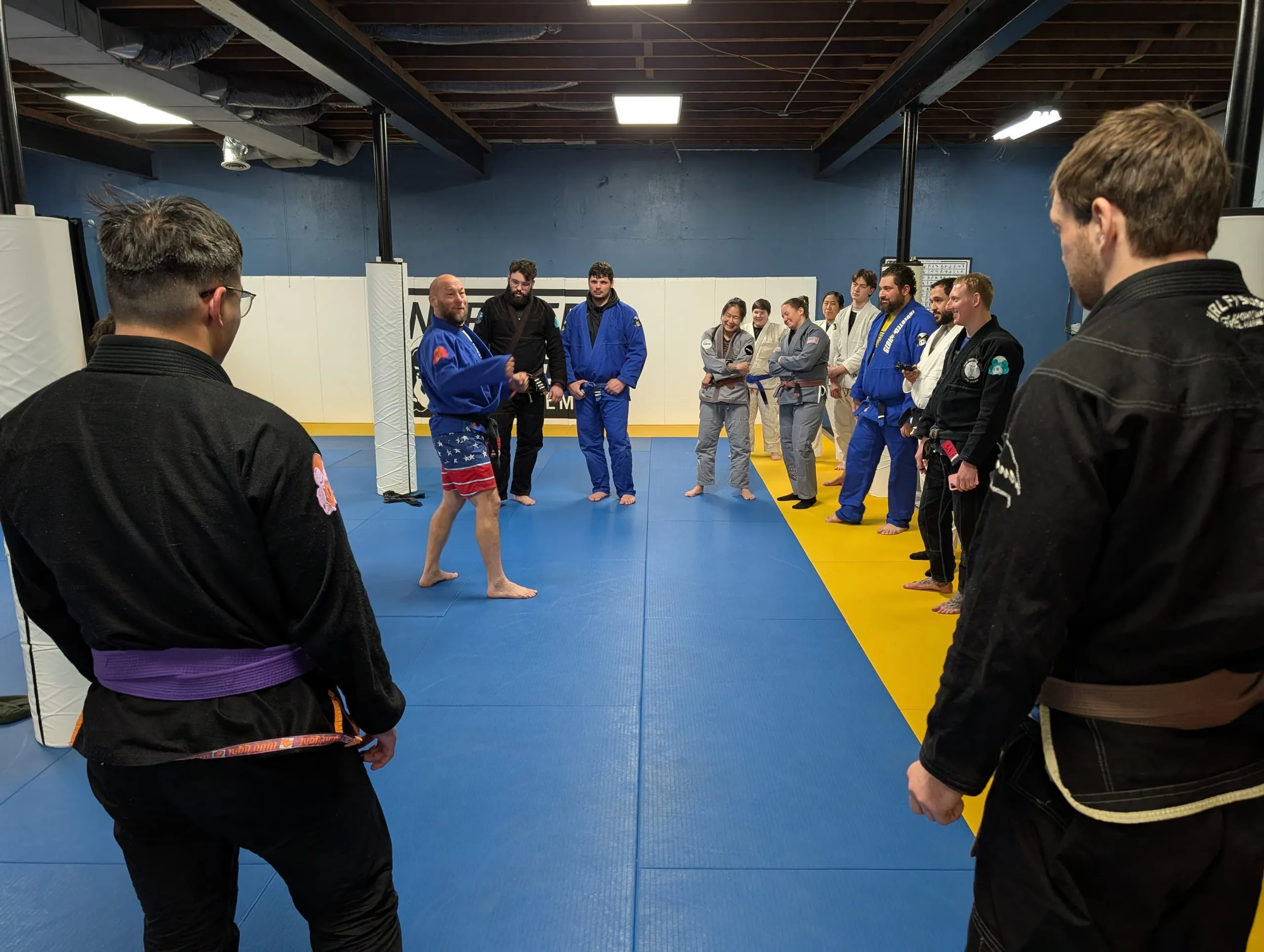 A jiu jitsu instructor addresses students gathered around the mats during a class.
