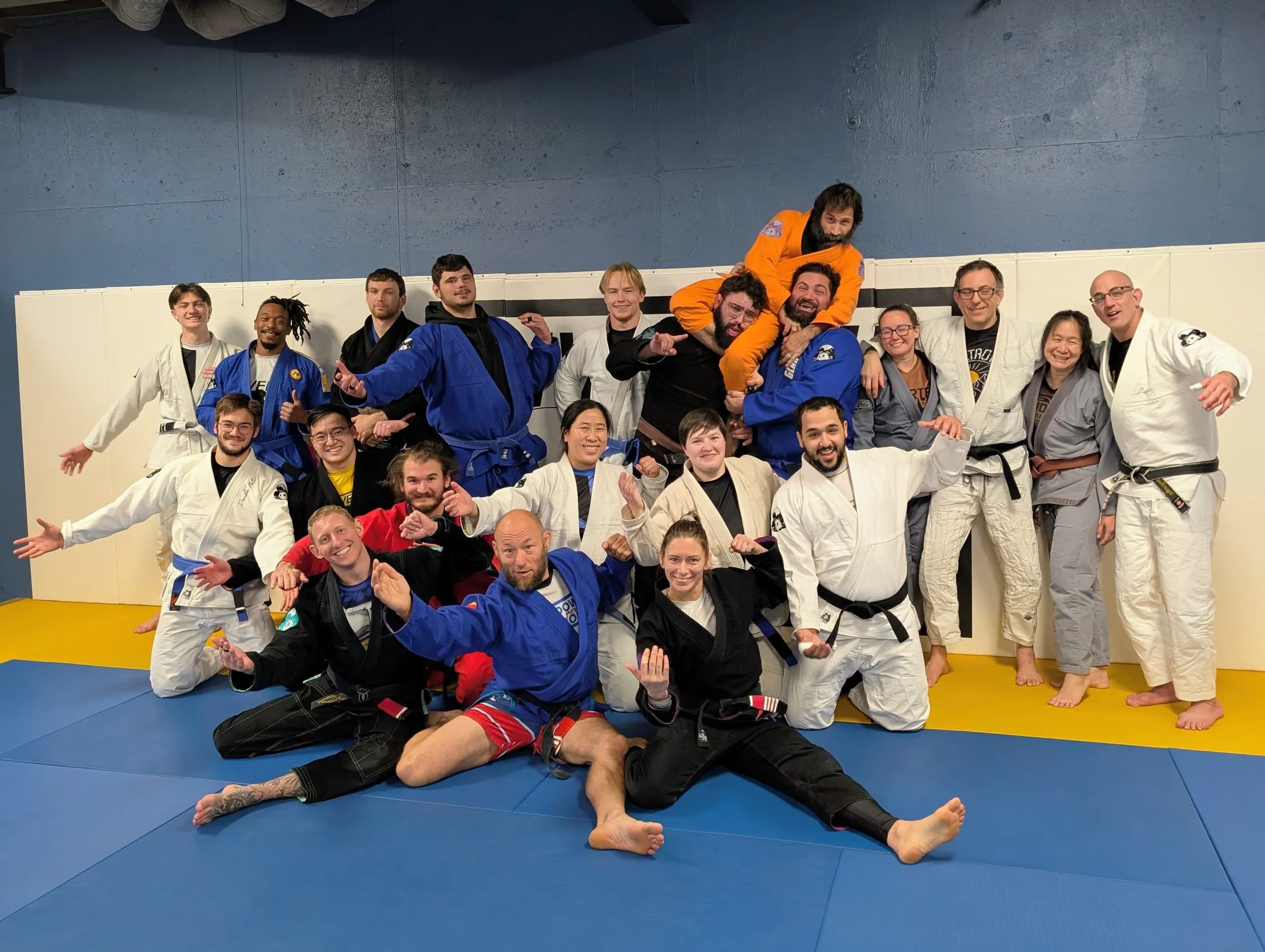 BJJ teammates in colorful gis laugh and strike playful poses for a group photo on blue and yellow mats.