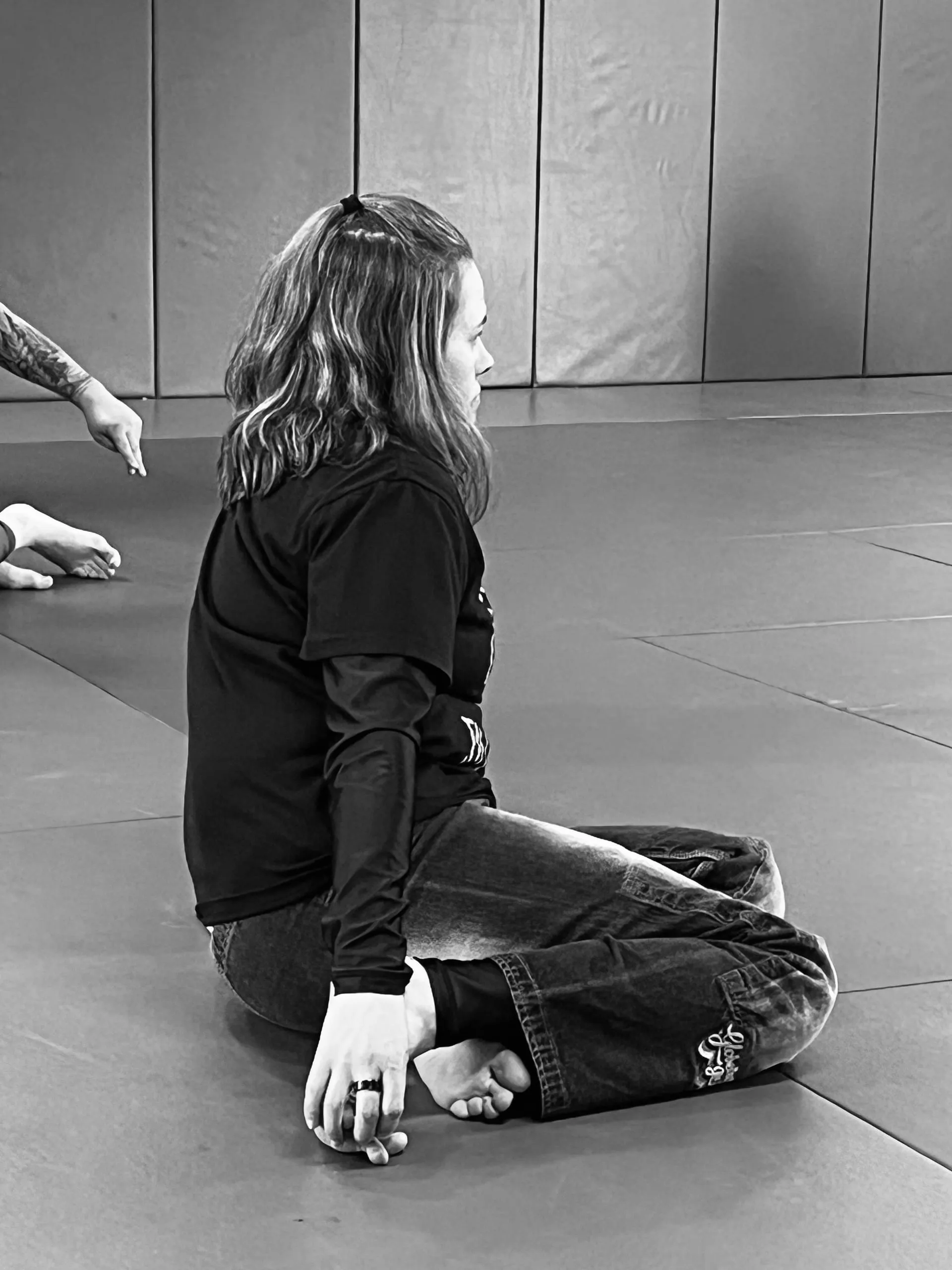 Black-and-white photo of Missy Baize seated on the mats in profile, watching a Brazilian Jiu Jitsu class.